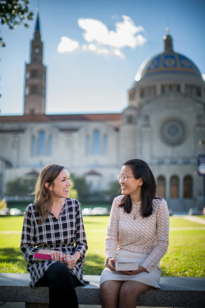 two female students sitting and talking in front of large school building